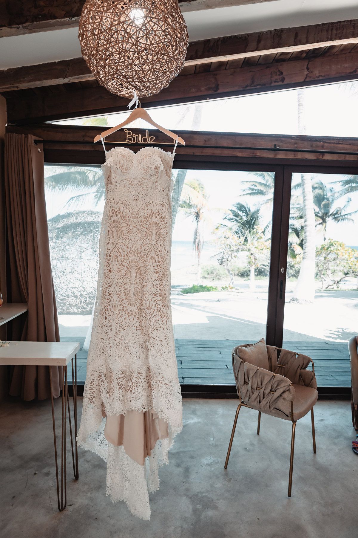 Elegant lace mermaid wedding dress hanging on a Bride hanger in a bright room with palm tree views.
