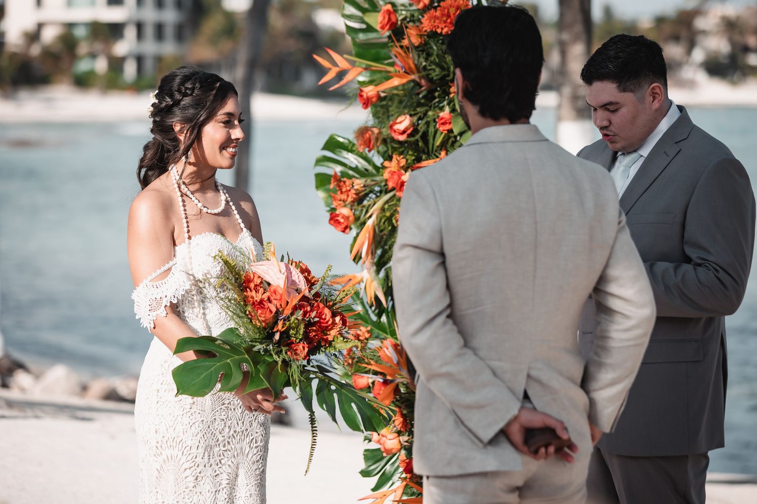 Bride holding tropical orange bouquet exchanges vows with groom at beachside wedding ceremony.