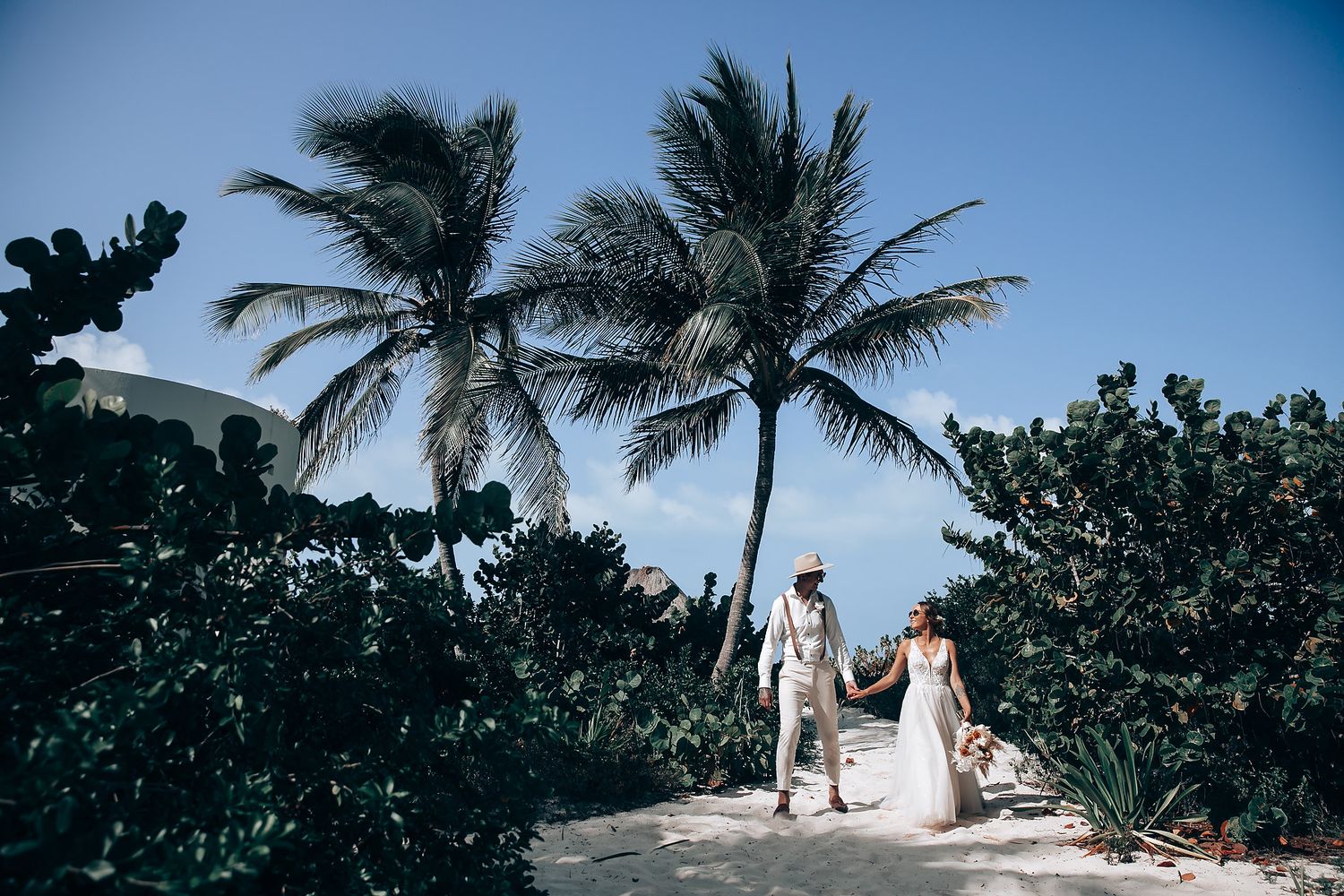 Tropical beach wedding scene with palm trees and white decor against blue skies.