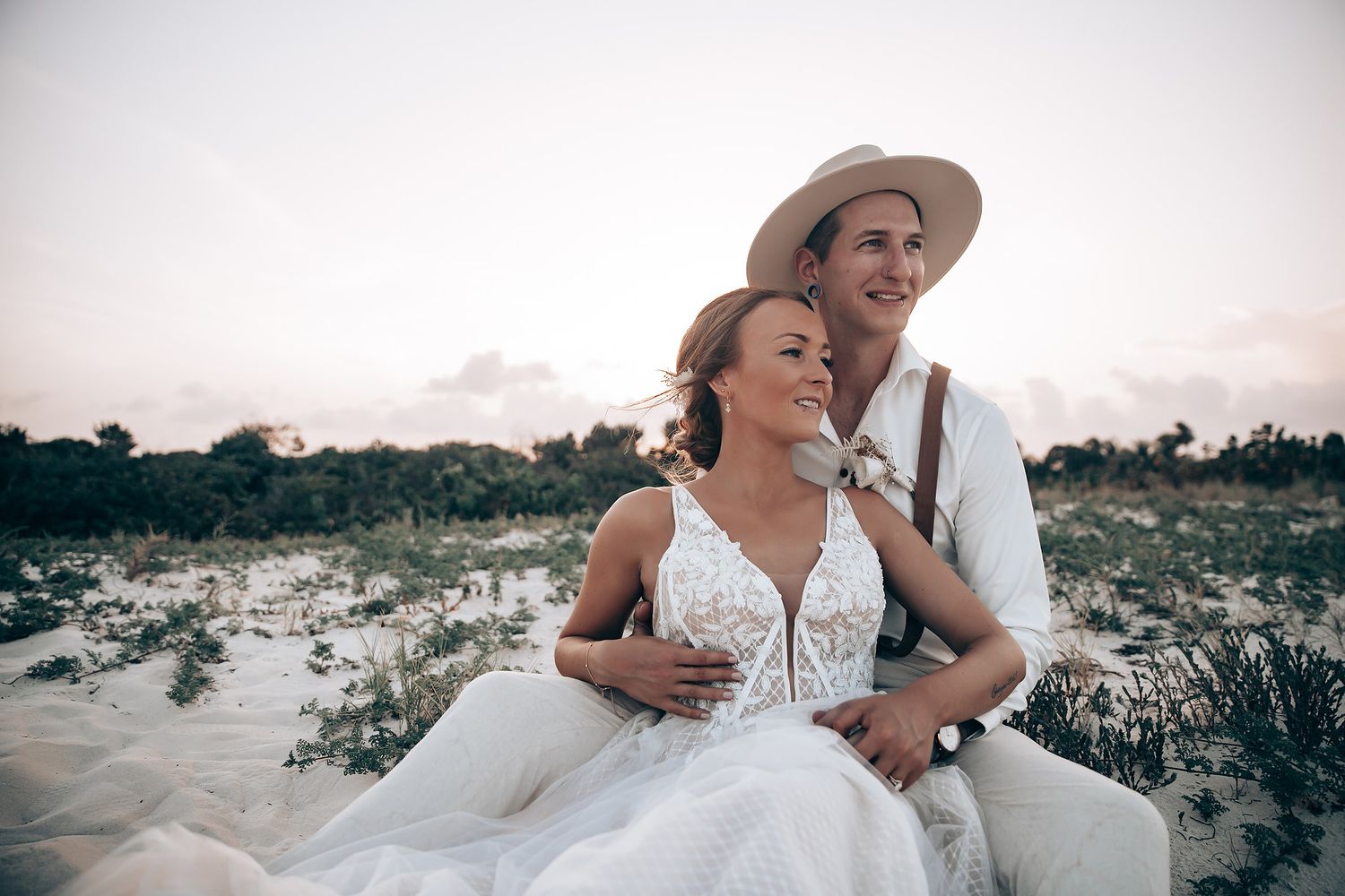 A tender moment between a couple in wedding attire sitting on sandy dunes at sunset.
