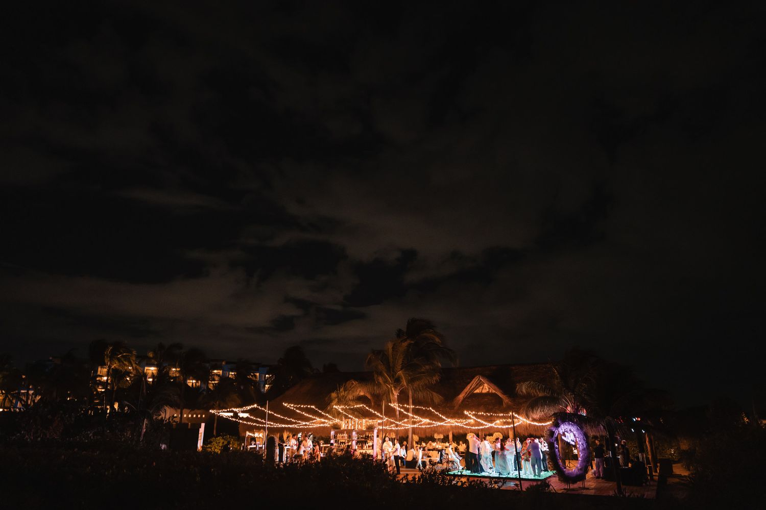 Festive outdoor wedding reception venue glowing with string lights under a dramatic cloudy night sky.