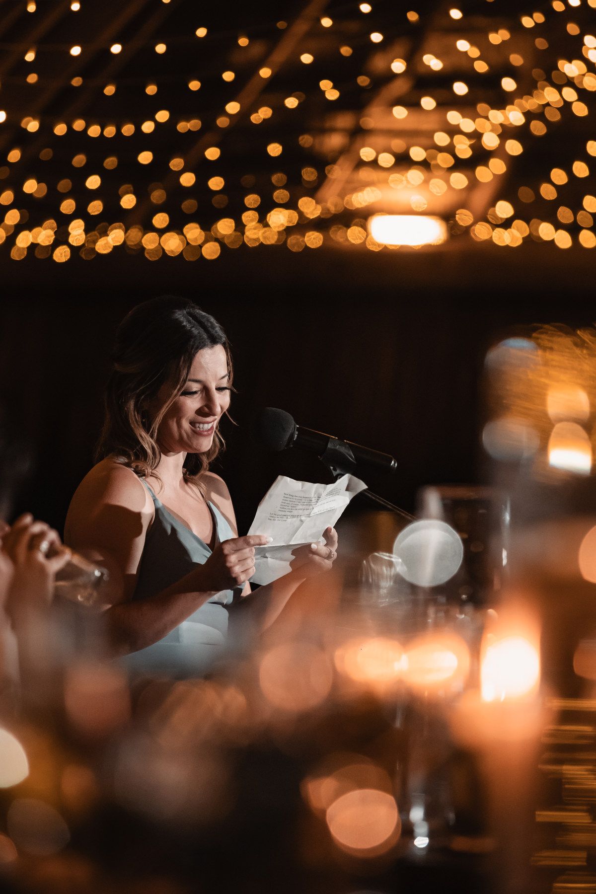 Smiling bridesmaid reading a wedding speech from paper under warm bokeh string lights at reception.