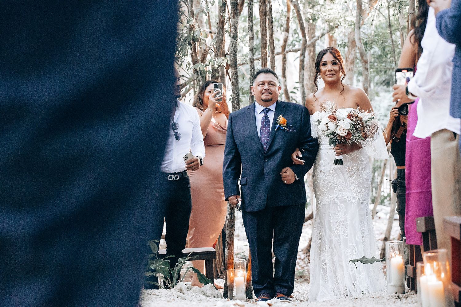 Outdoor wedding ceremony in a wooded setting with guests seated on white chairs.