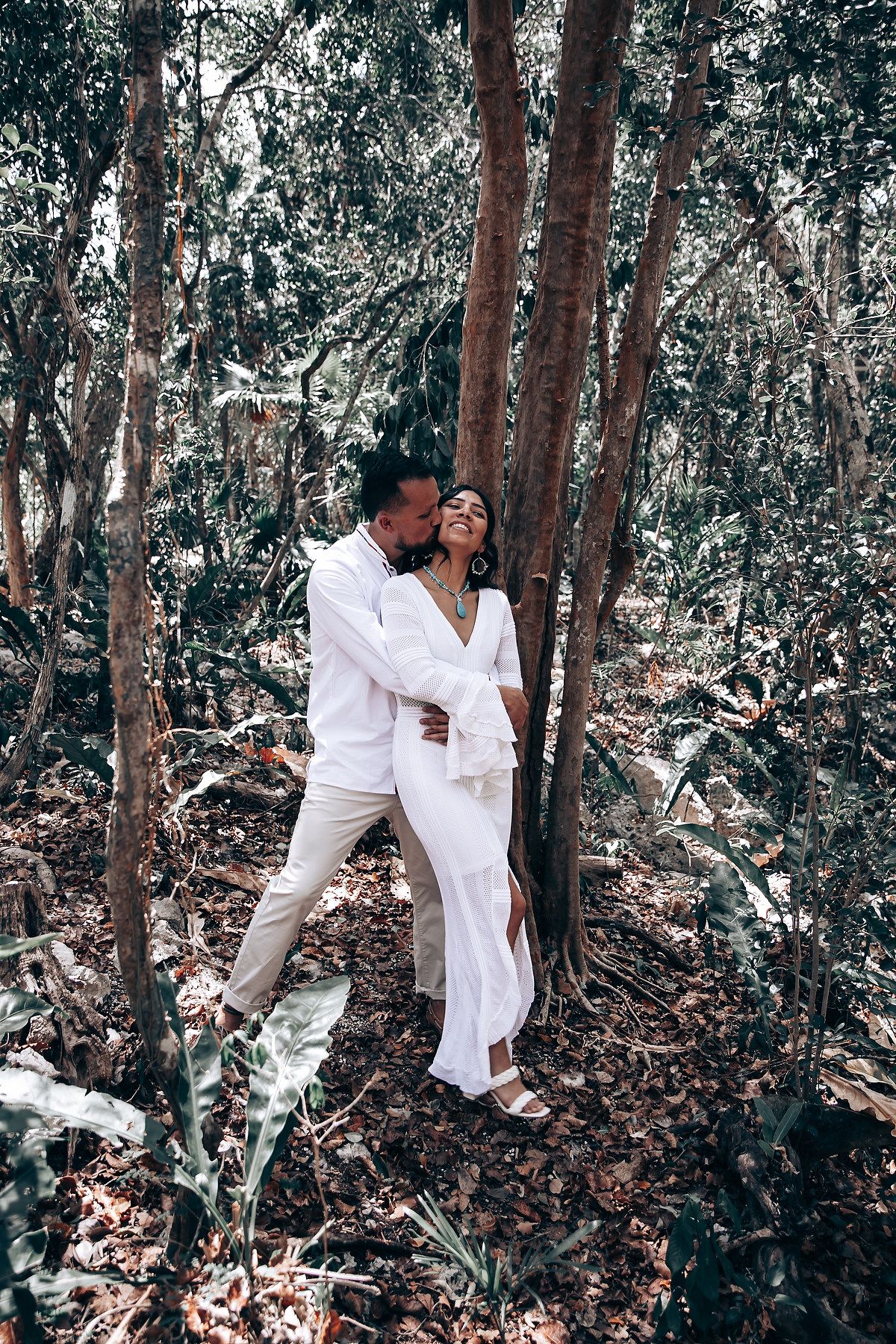 Couple in white outfits sharing an intimate moment in a forest setting.