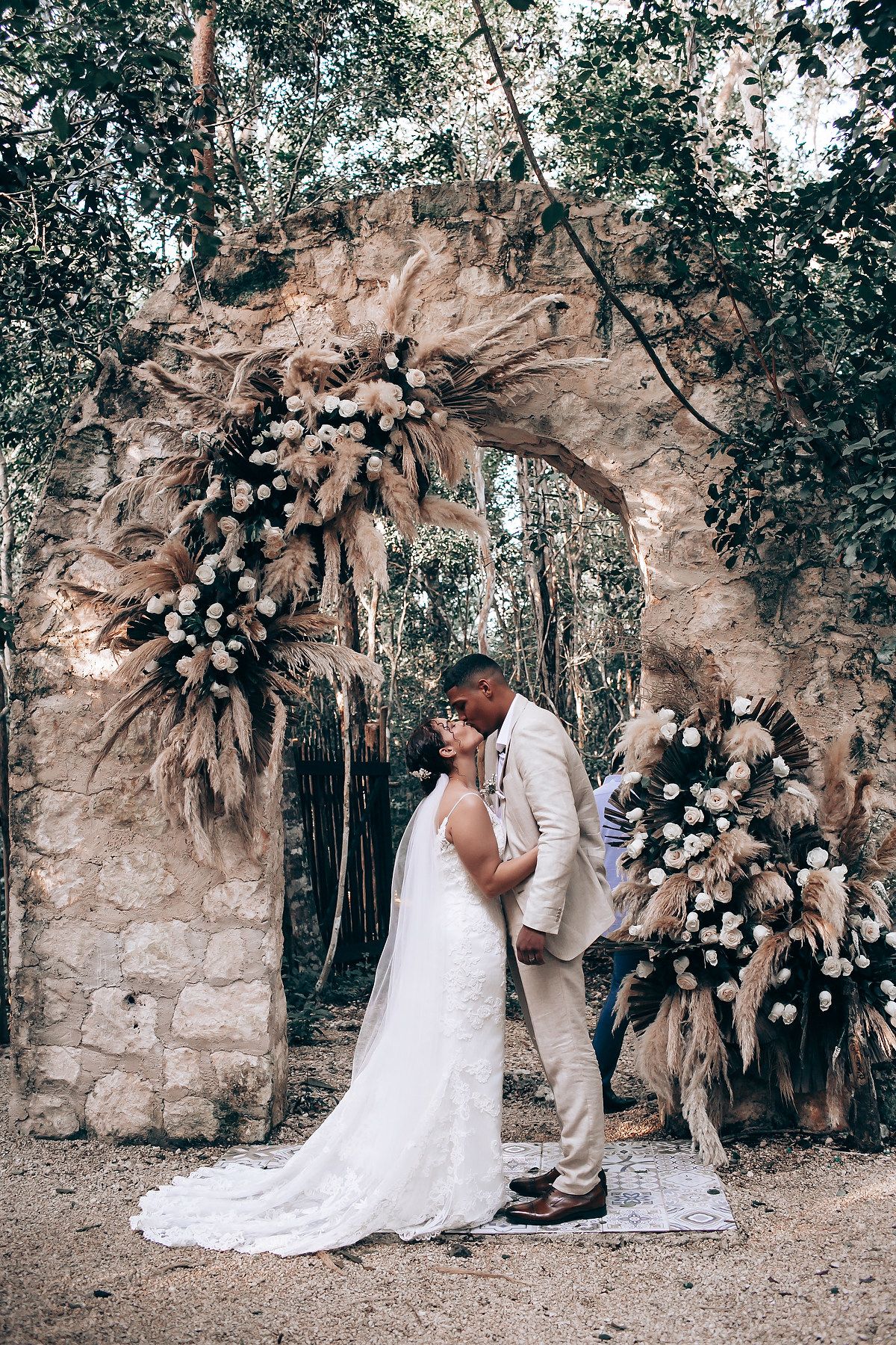 A romantic wedding ceremony set against a stone archway decorated with white flowers in a forest setting.