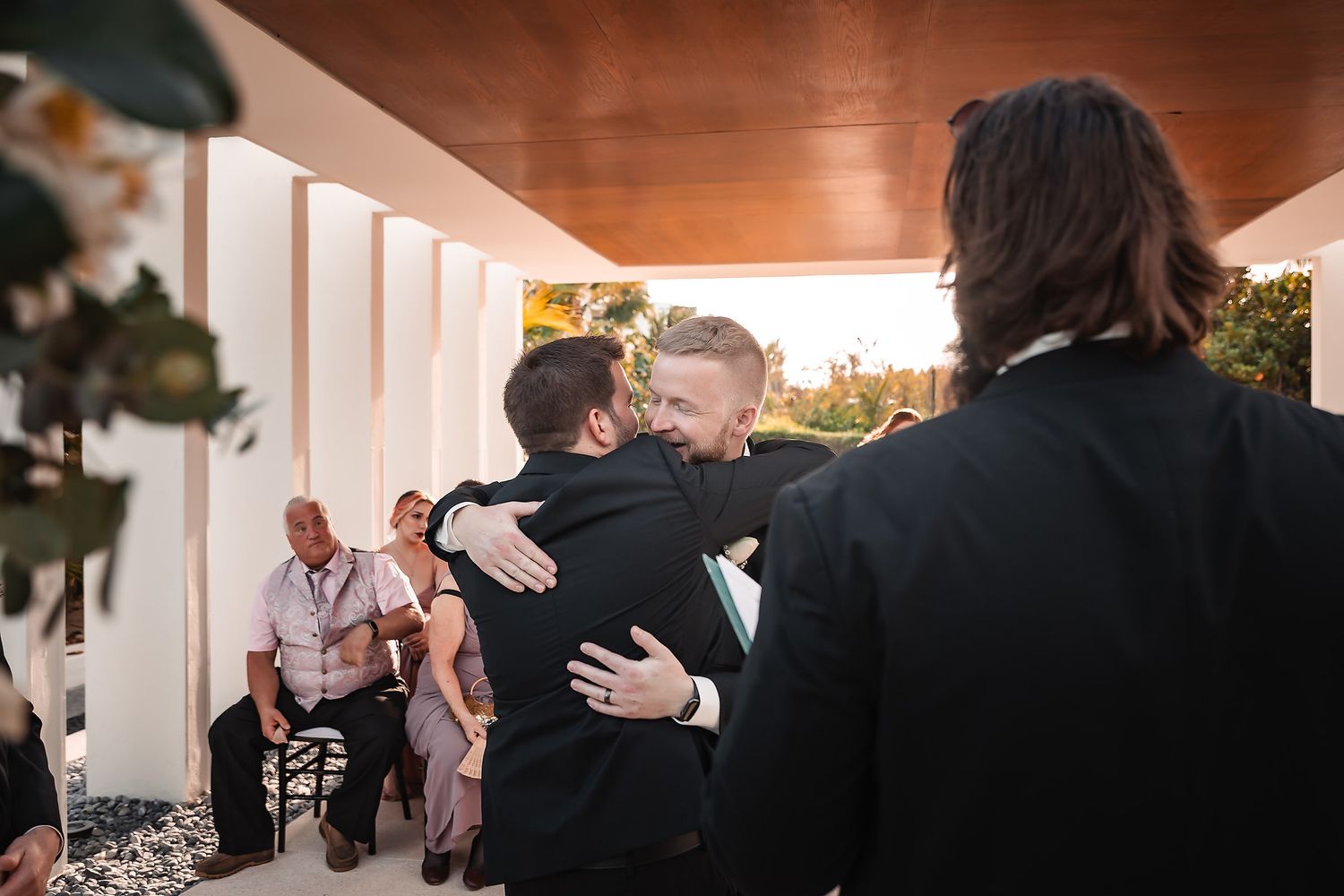 Two grooms share an emotional hug during their wedding ceremony outdoors, with guests seated in the background.