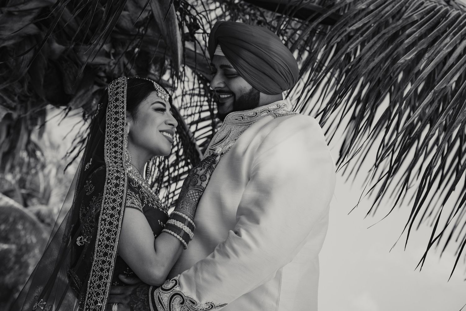 Black and white portrait of a Sikh bride and groom sharing a joyful moment under tropical palm trees.