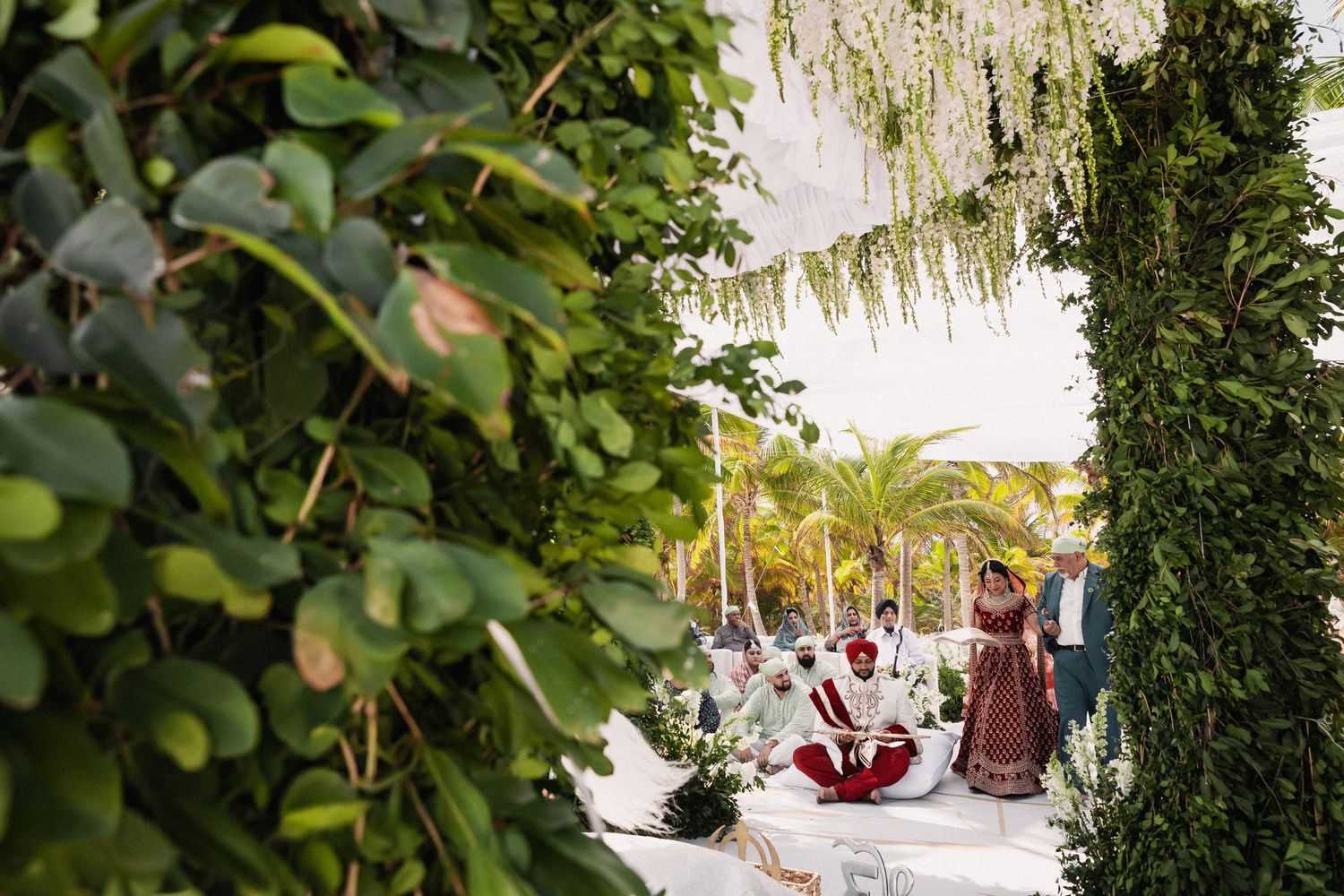 Outdoor Sikh wedding ceremony framed by lush greenery with guests seated under floral canopy.