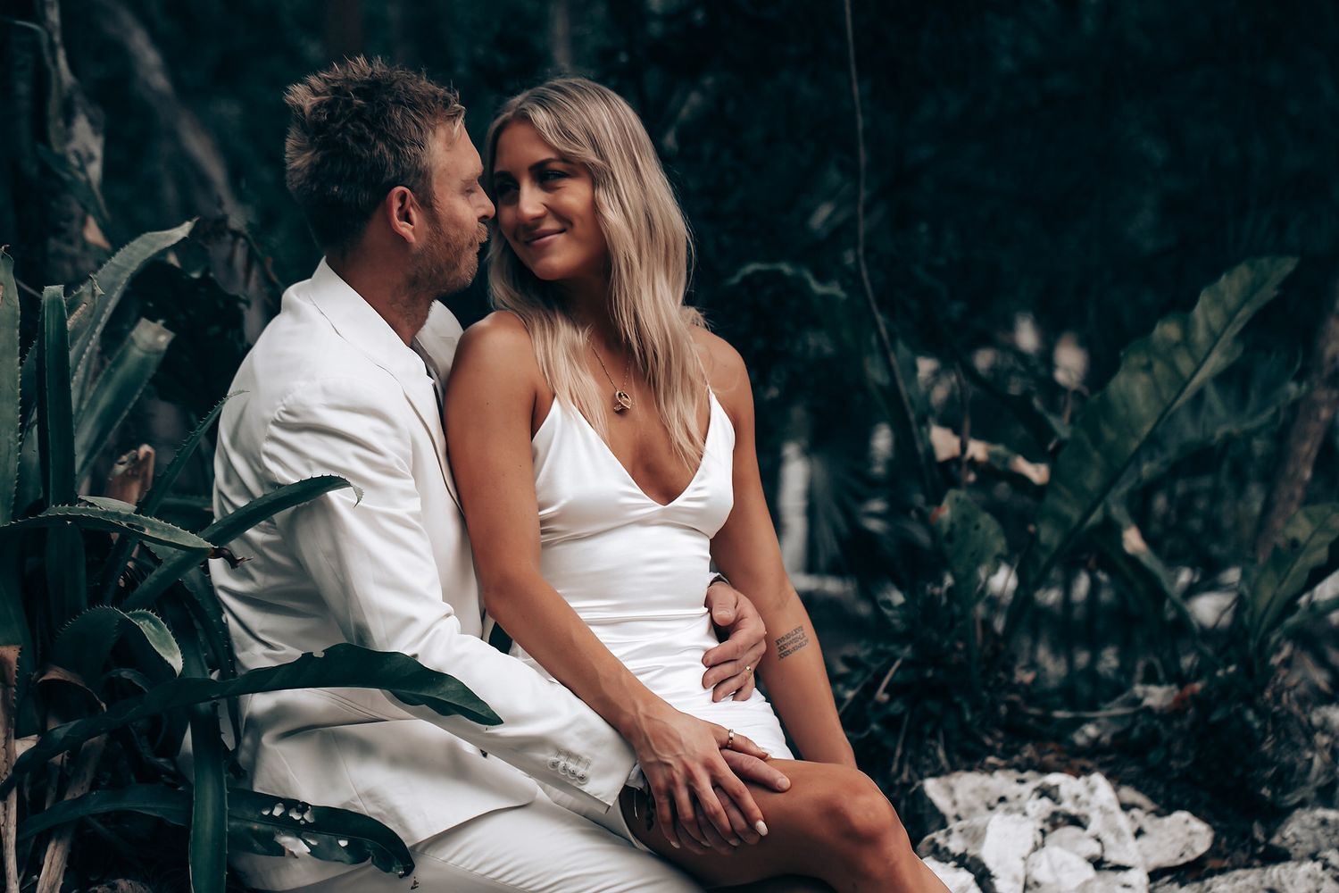 A romantic couple in white attire sharing an intimate moment in a lush tropical setting.