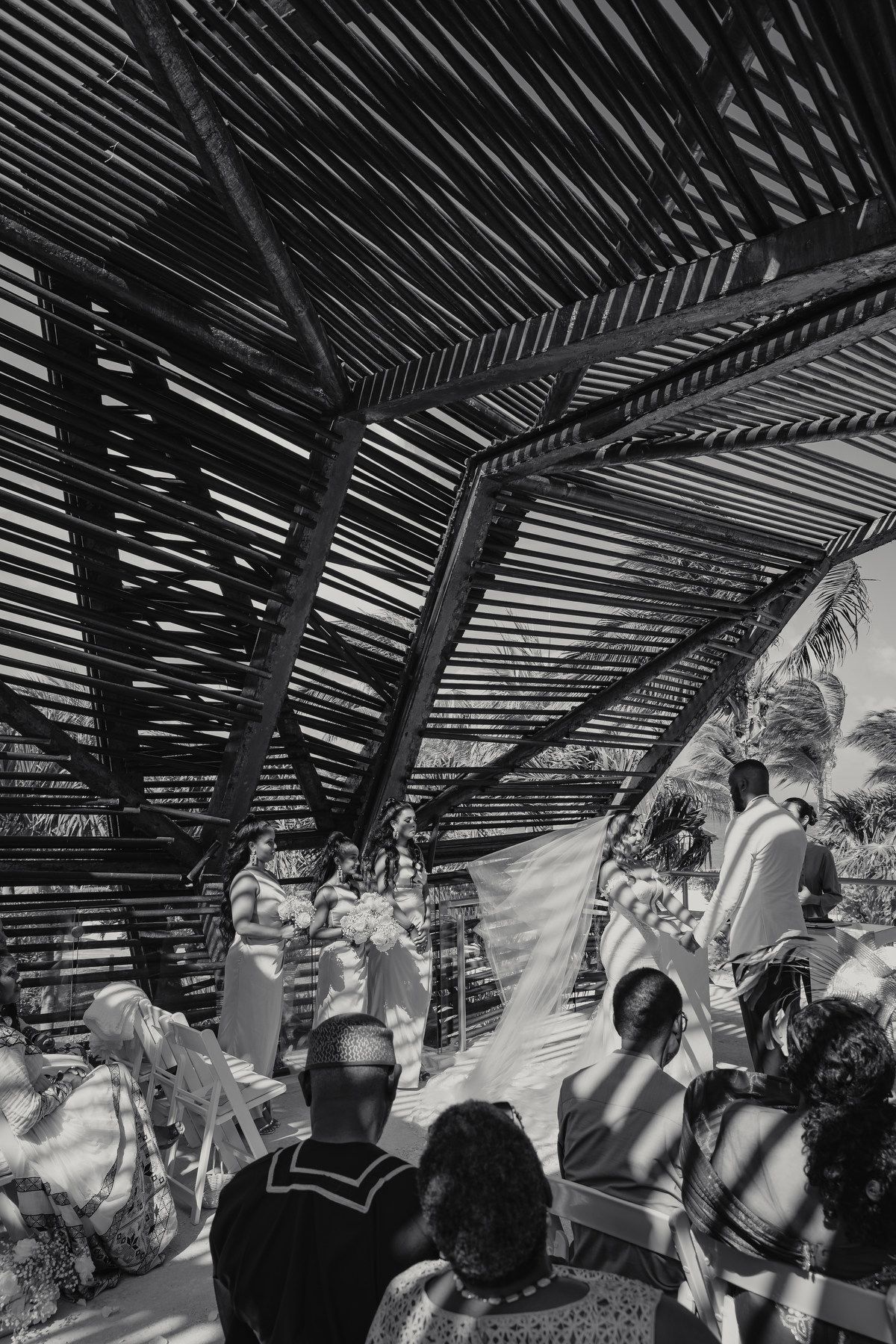 Black and white photo of outdoor wedding ceremony under bamboo pergola with seated guests.