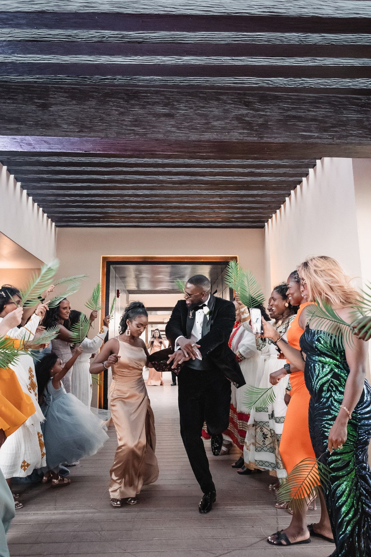 Bride and groom dance through a joyful wedding reception exit tunnel with guests waving tropical palm leaves.