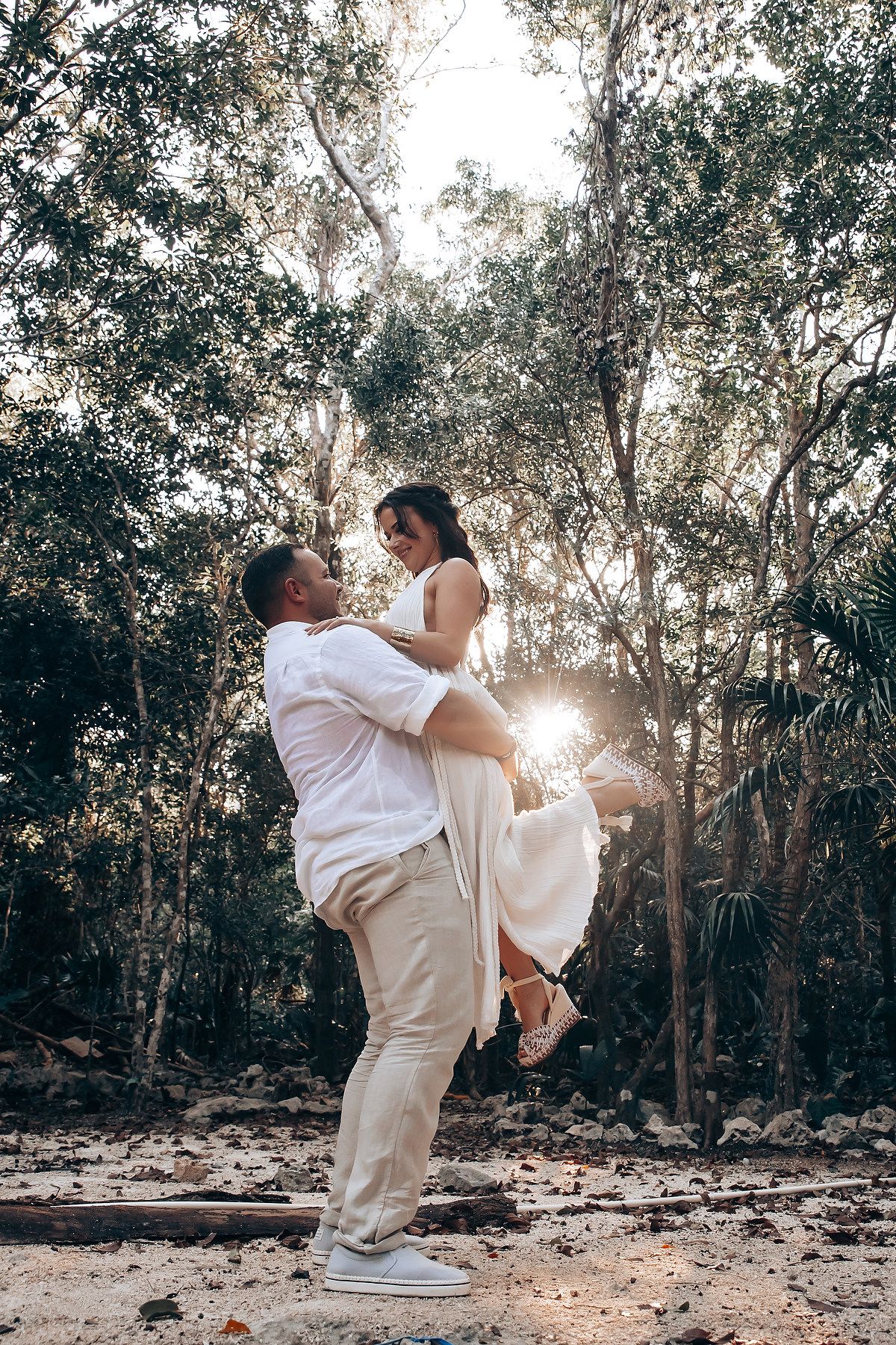 A couple in white outfits performs a dance lift sequence in a sunlit wooded area with trees in the background.