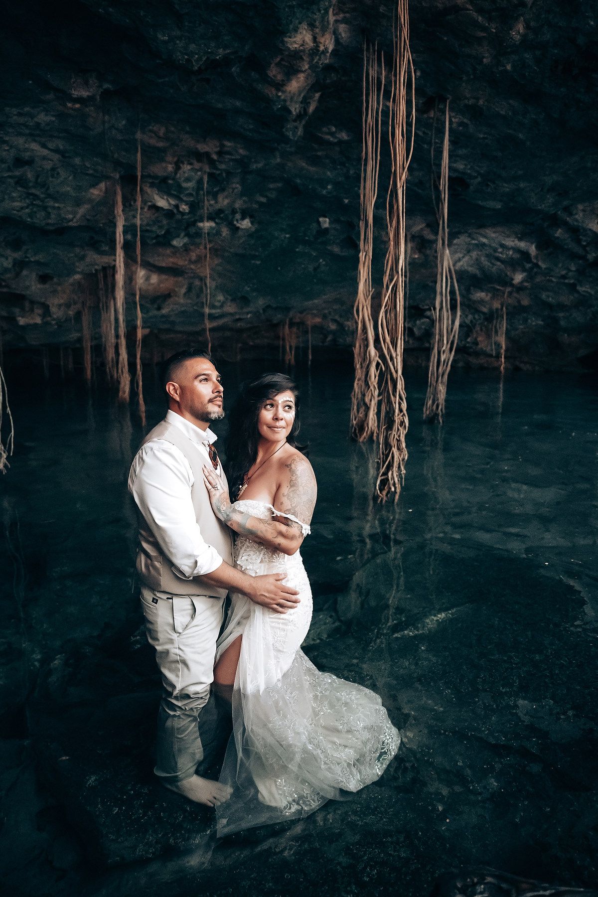 A couple embraces in an atmospheric cave setting with dramatic lighting and hanging vines.