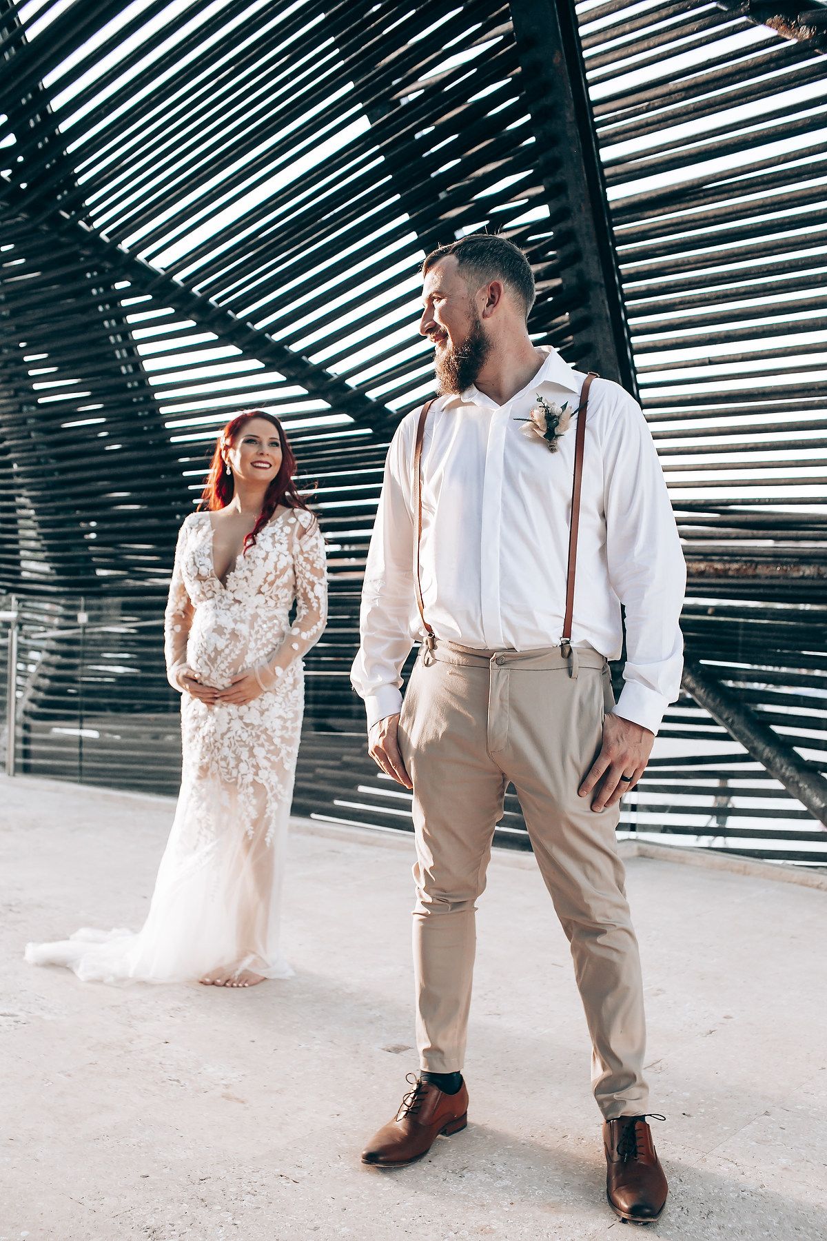 Romantic couple in wedding attire posing under geometric wooden structure with dramatic lighting.