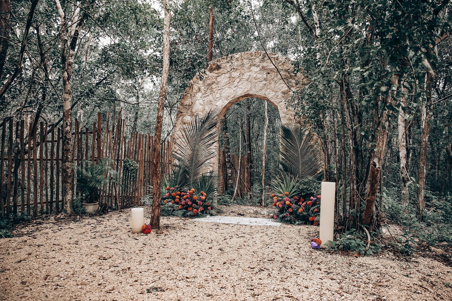 A rustic stone archway entrance along a dirt path in a wooded area with decorative red accents.