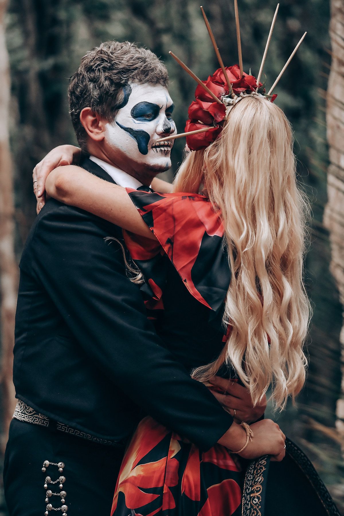 Gothic couple in Day of the Dead makeup and costumes embrace romantically in a moody forest setting.
