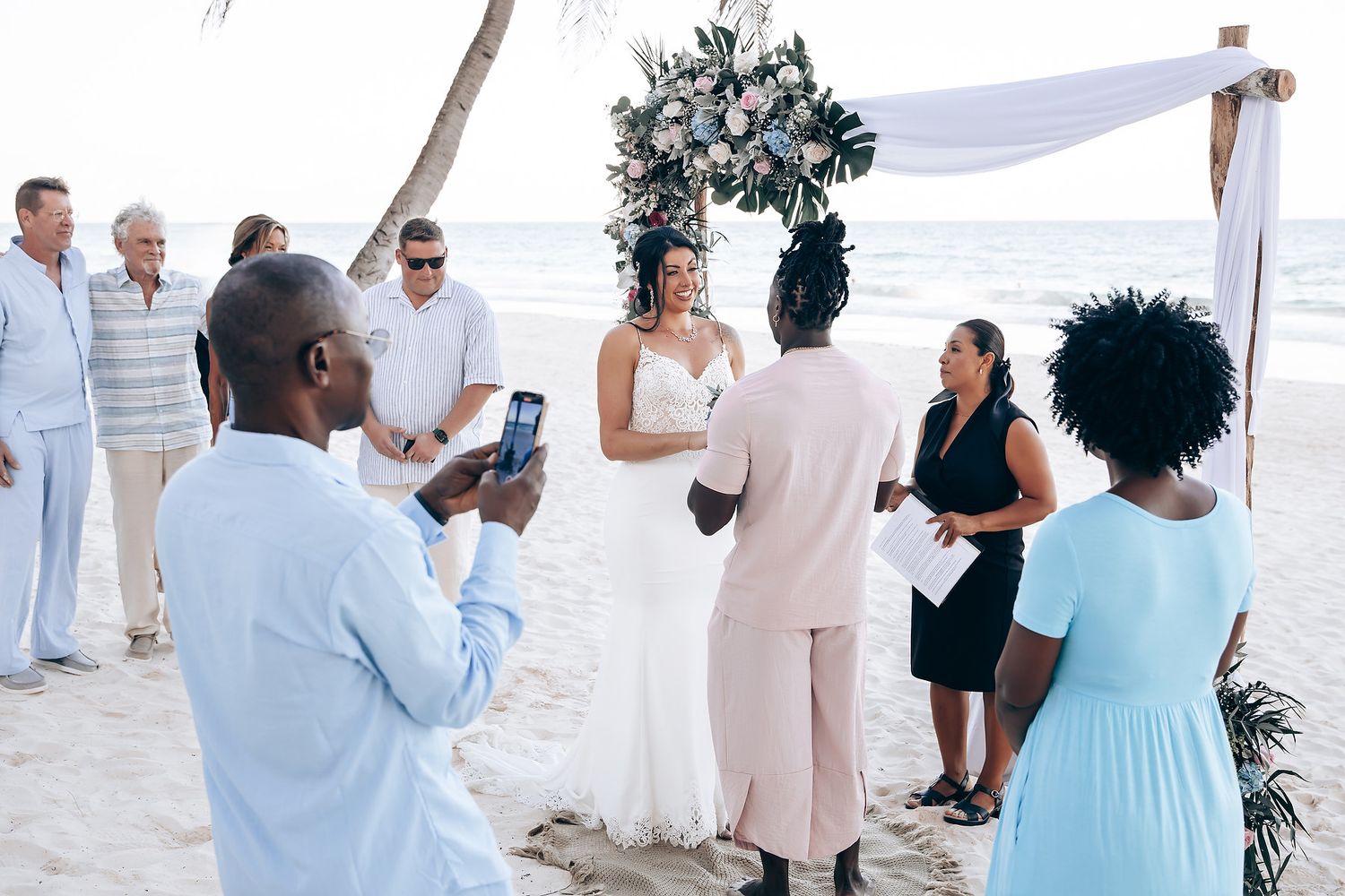 A wedding ceremony taking place on a beach, with a floral arch and guests in attendance.