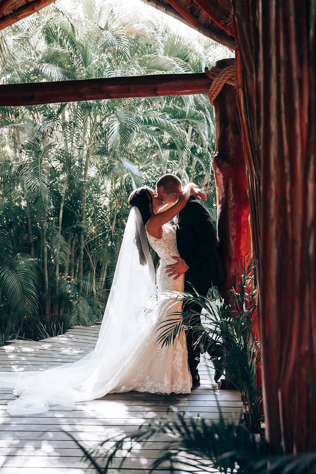 A romantic wedding couple embraces in a tropical garden setting framed by red curtains and palm trees.