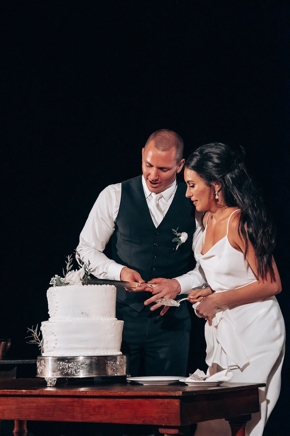 Couple shares a joyful moment while cutting their white wedding cake against dark background.