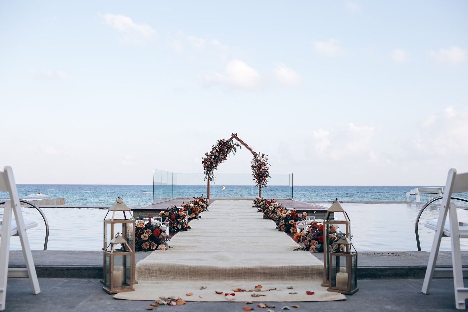 Beachfront wedding ceremony setup with white chairs and floral arch overlooking the ocean.