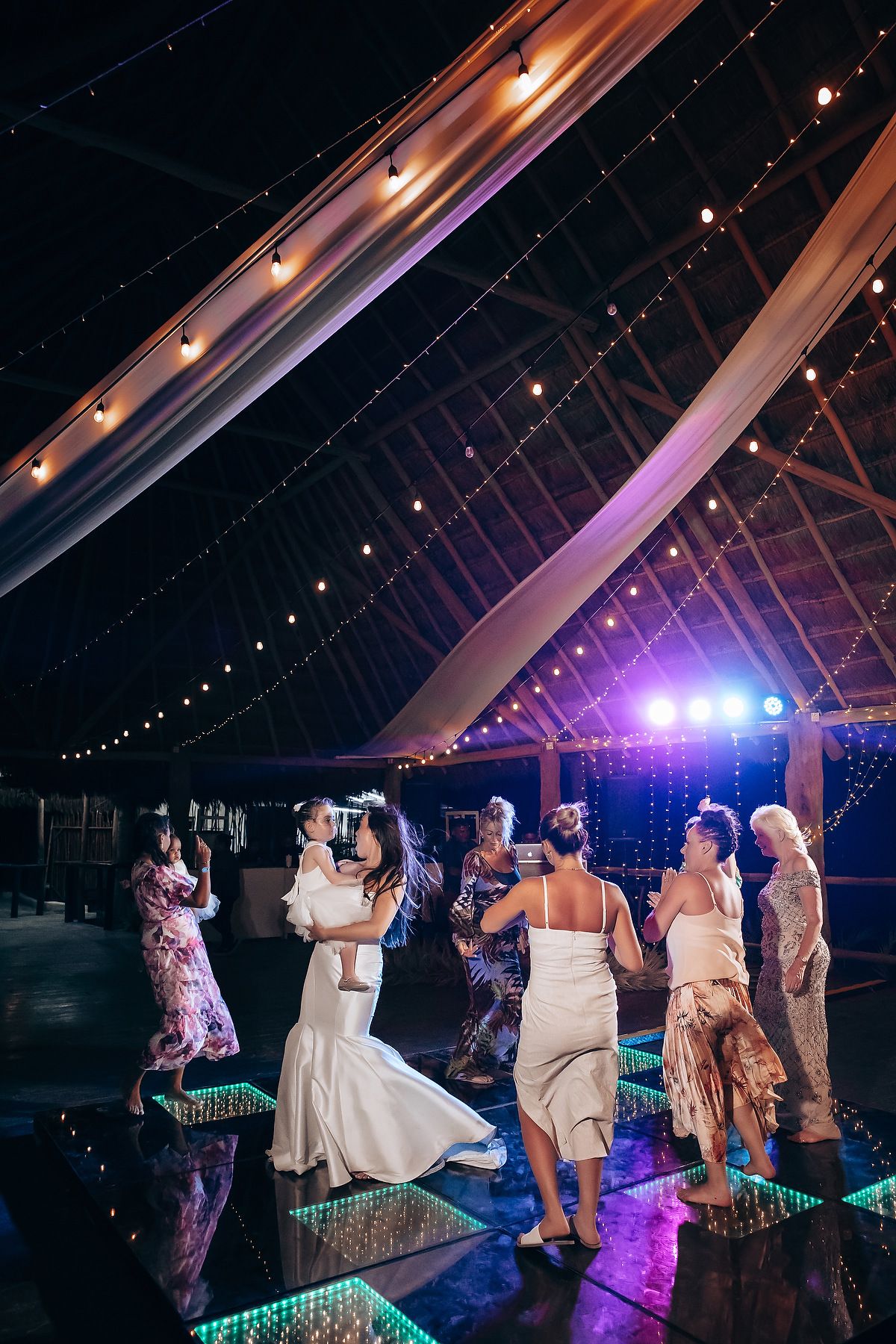 Wedding guests dance under colorful lights on an illuminated dance floor during an evening reception celebration.