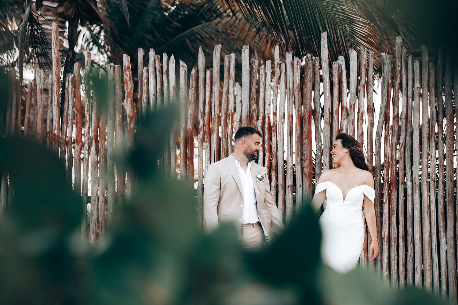 Couple in wedding attire walking along rustic wooden fence in tropical beach setting at sunset.