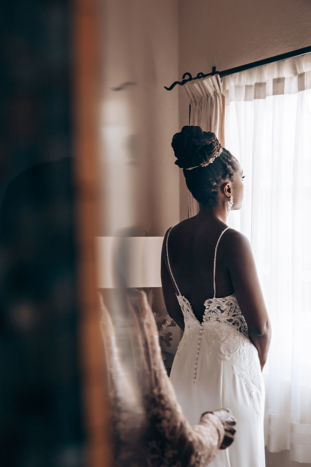 Elegant bride in white dress photographed from behind near a sunlit window.