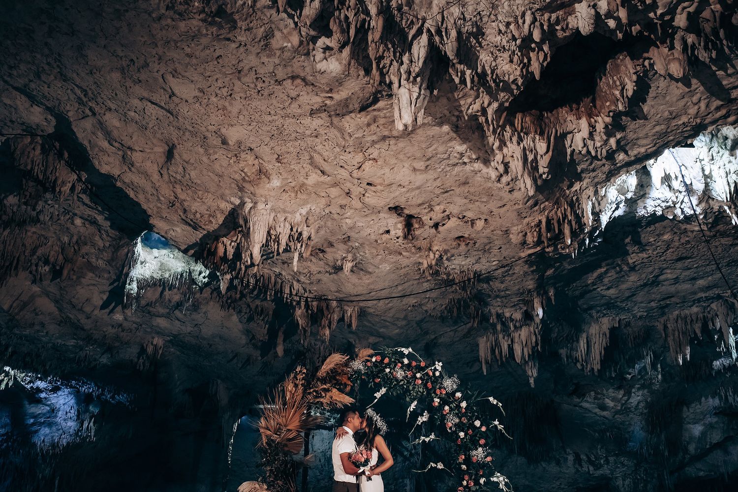 Couple embraces under an illuminated floral arch inside a dramatic cave with stalactites and natural rock formations.