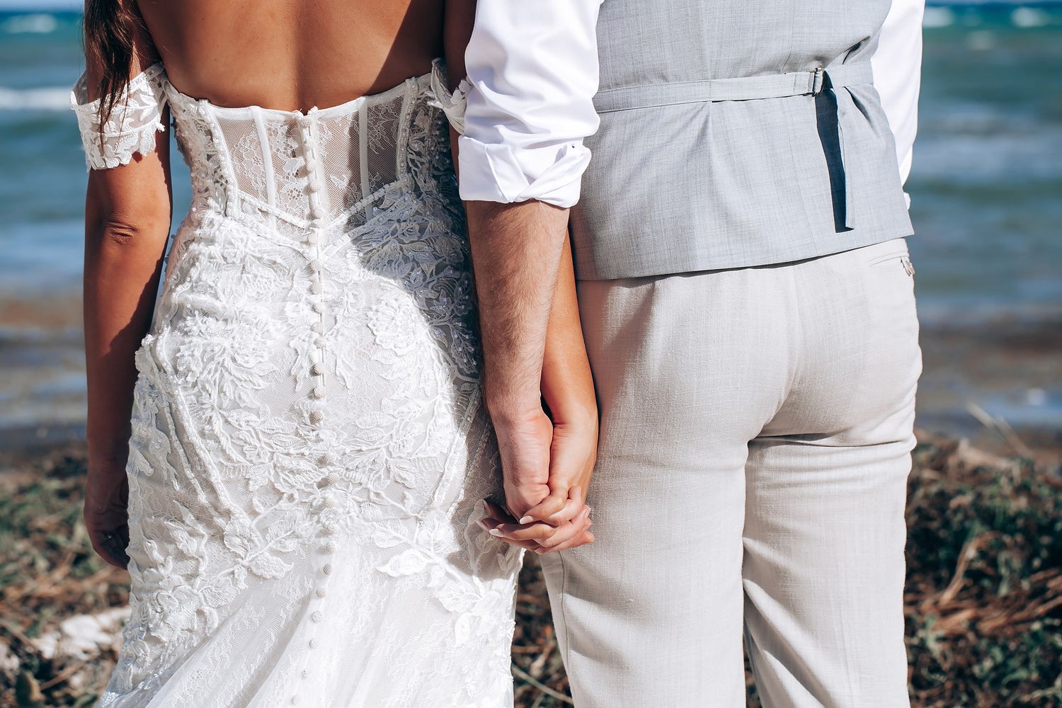 Close-up of bride's lace dress detail and groom's white attire against ocean backdrop.