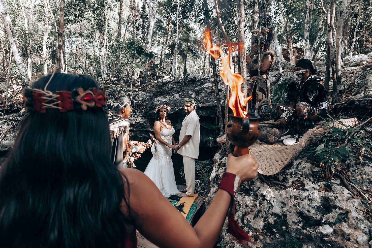 A wedding ceremony takes place in a scenic wooded forest setting with natural stone formations and sunlight filtering through trees.