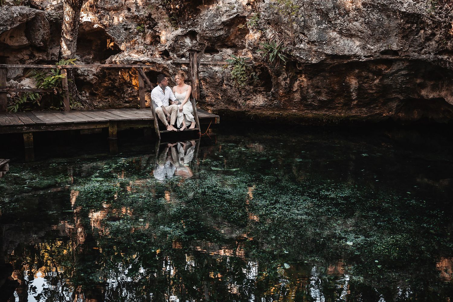 Couple embracing on a wooden dock over a stunning emerald cenote surrounded by rocky walls in Mexico.