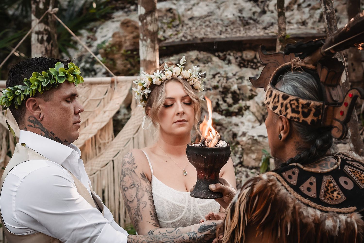 Tattooed couple in bohemian wedding attire during ritual ceremony with flame torch and tribal officiant outdoors.