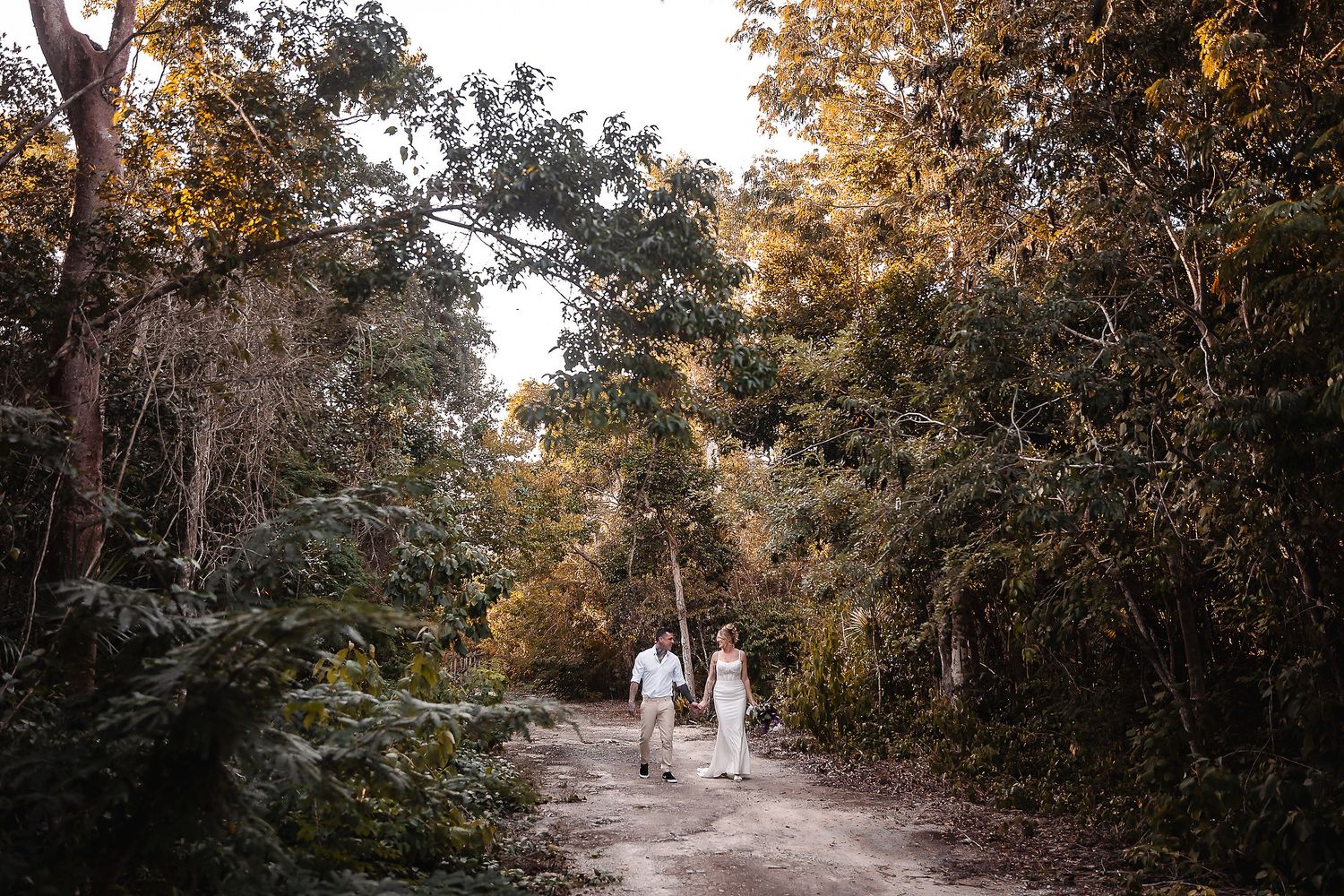 Couple walking hand in hand on a forest path surrounded by lush green and golden trees.