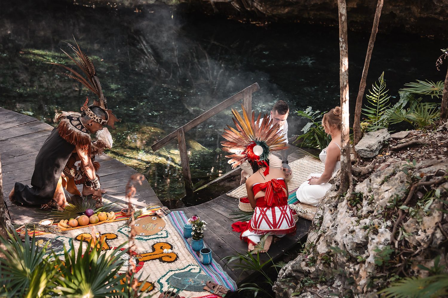 Traditional Mayan ceremonial performers in colorful costumes near a cenote surrounded by lush tropical vegetation.