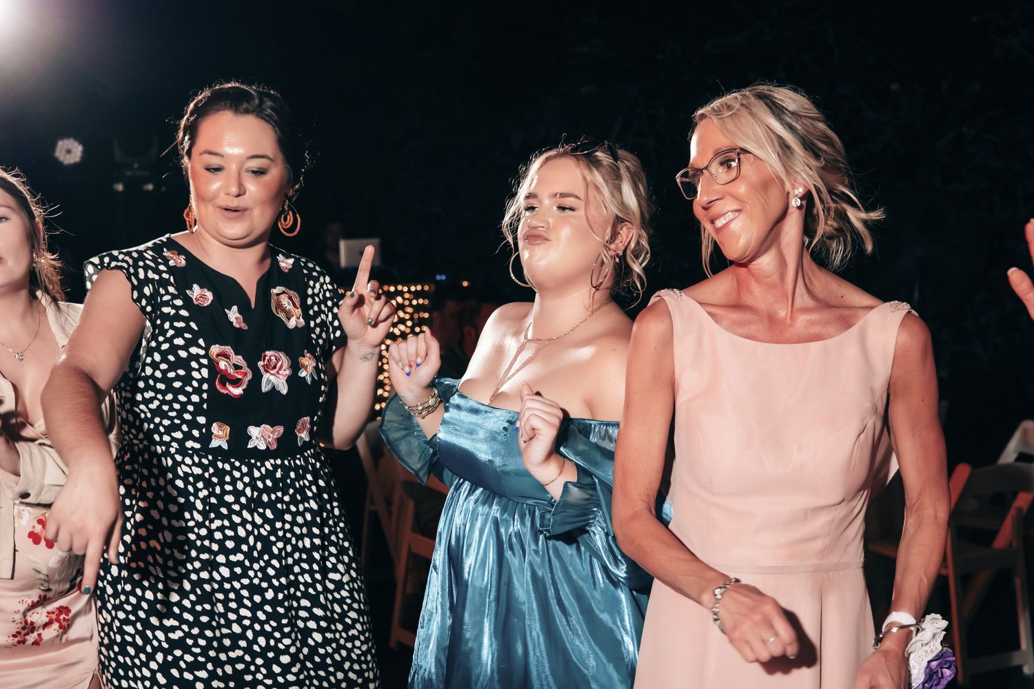 Three women in elegant dresses dancing and celebrating joyfully at an outdoor evening wedding reception.