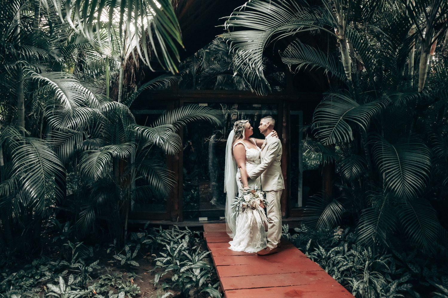 Bride and groom embrace on a red bridge surrounded by lush tropical greenery in a romantic outdoor wedding setting at Dreams Tulum