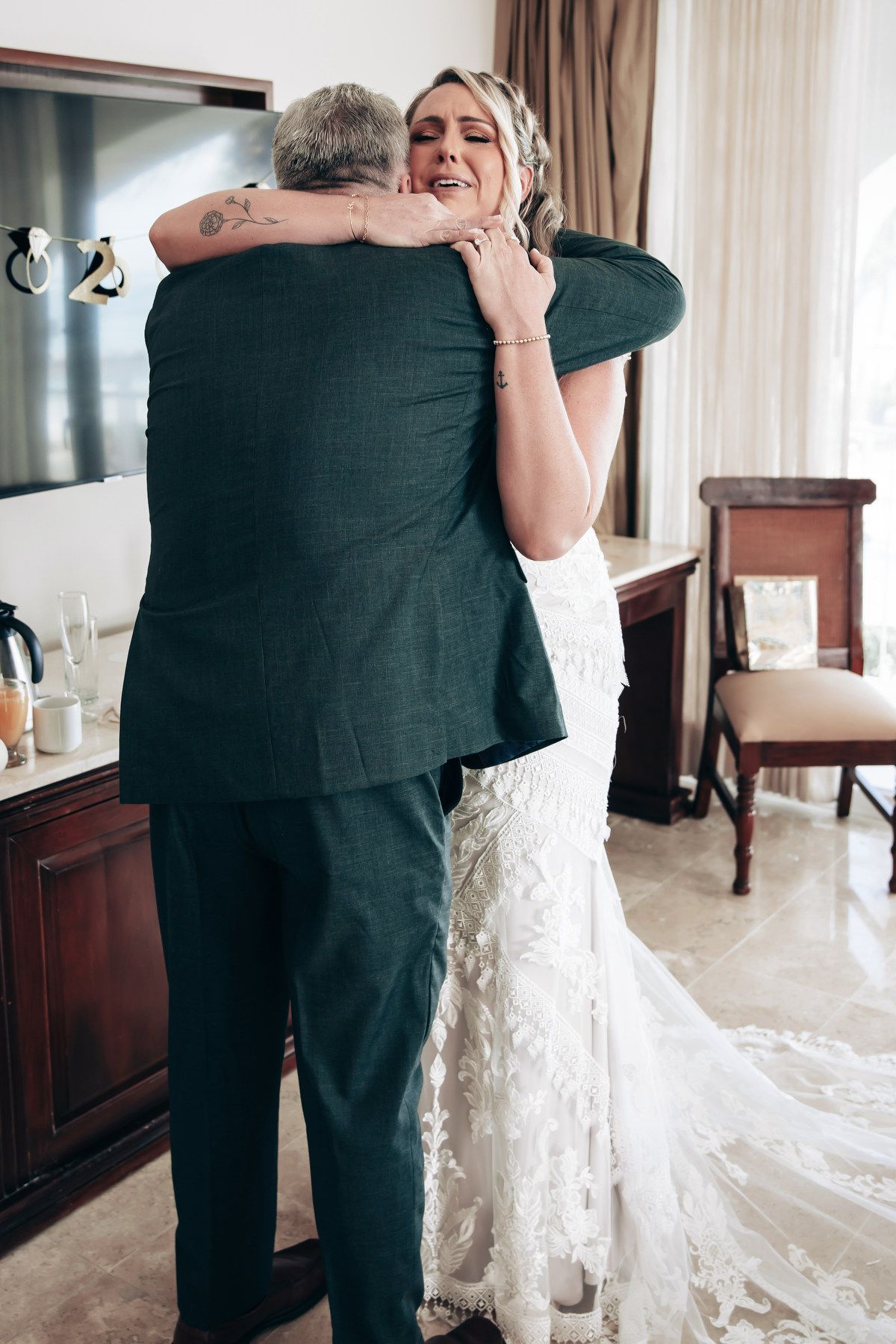 Emotional bride in lace wedding gown hugging a man in a dark suit during a heartfelt first look moment indoors.