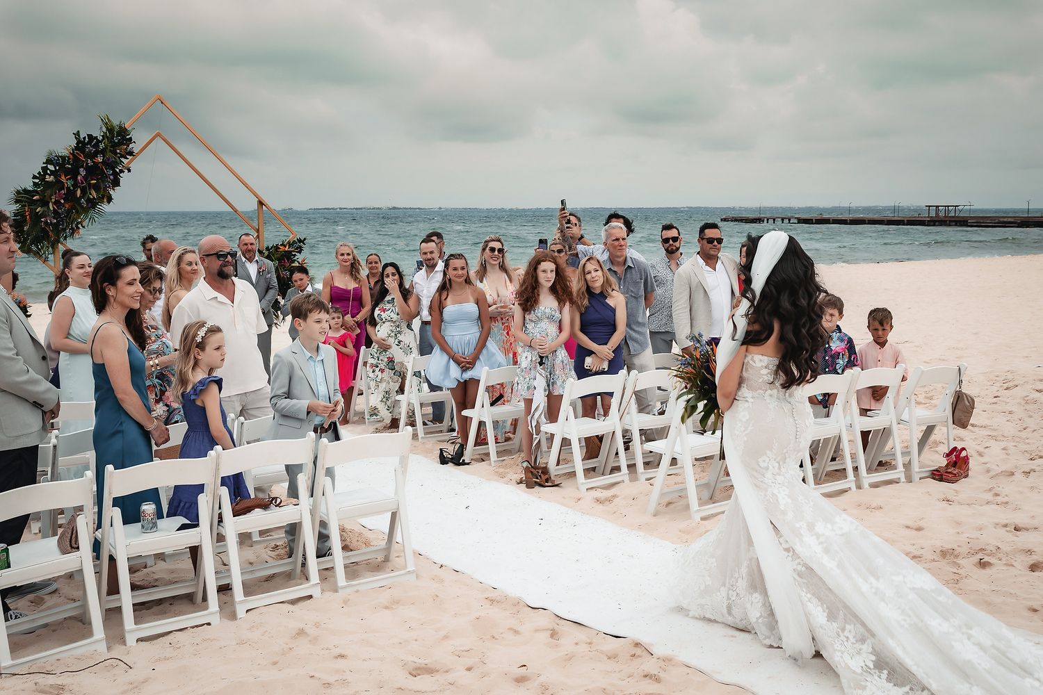 Beach wedding ceremony with guests seated in white chairs, bride in flowing gown, ocean backdrop, cloudy sky.