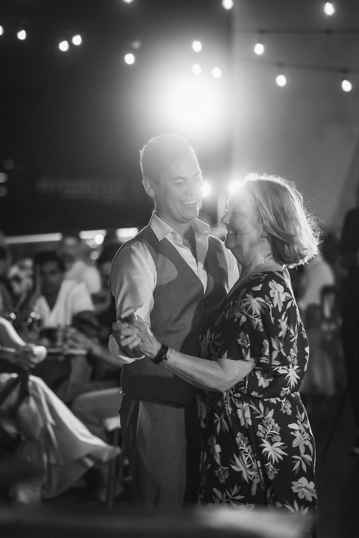 Black and white photo of a couple sharing a romantic dance under string lights at an elegant evening event.
