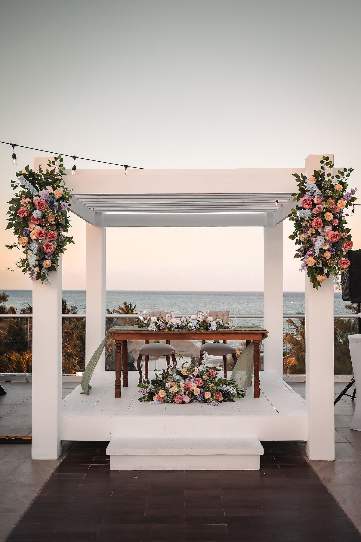 Elegant beachfront wedding altar with white pergola, pink floral arrangements, and wooden sweetheart table at sunset.