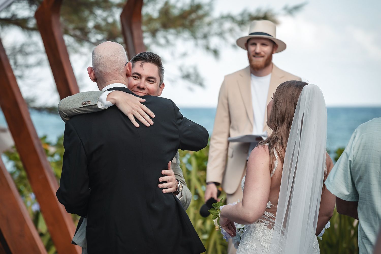 Groom hugs guest at outdoor wedding ceremony while bride stands nearby in white gown.