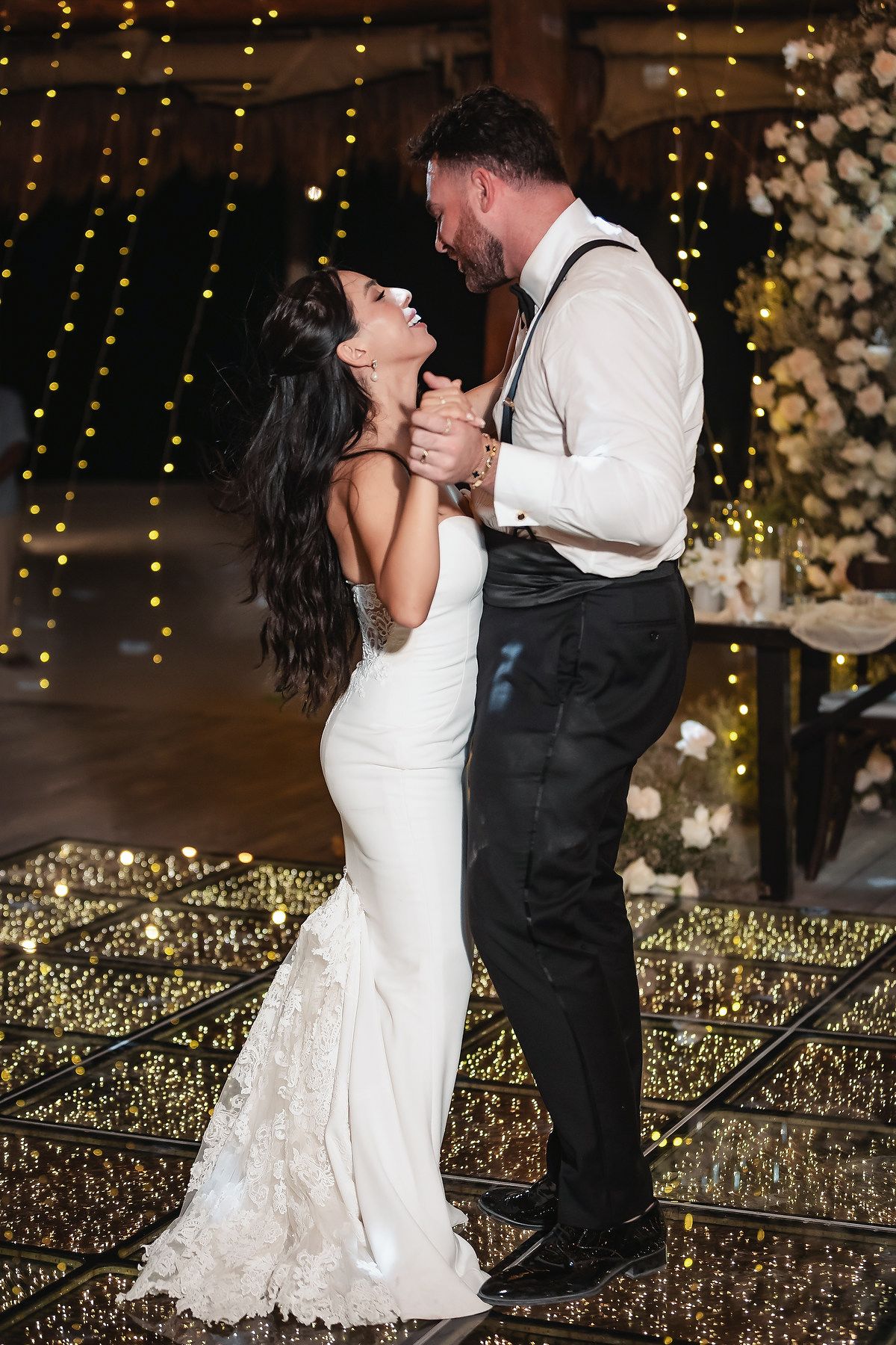 Bride and groom share first dance on glittering mirror floor surrounded by warm fairy lights at elegant wedding reception.