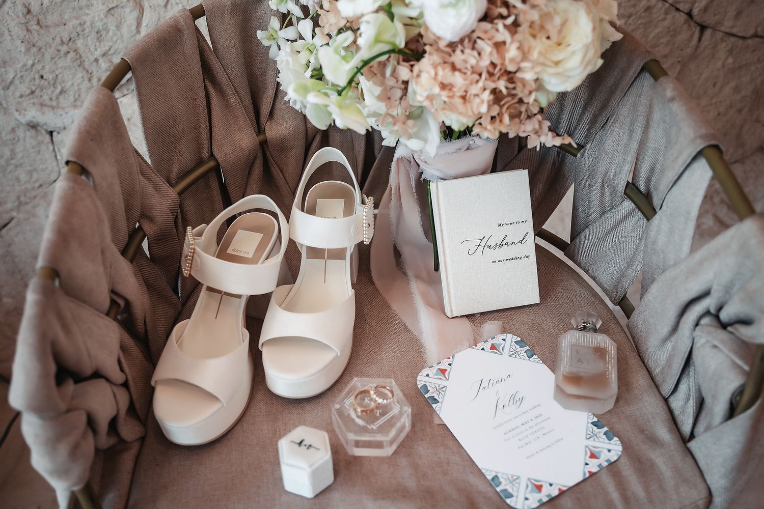 Elegant wedding flat lay with white block heel sandals, floral bouquet, invitation cards, and perfume bottle on blush fabric.