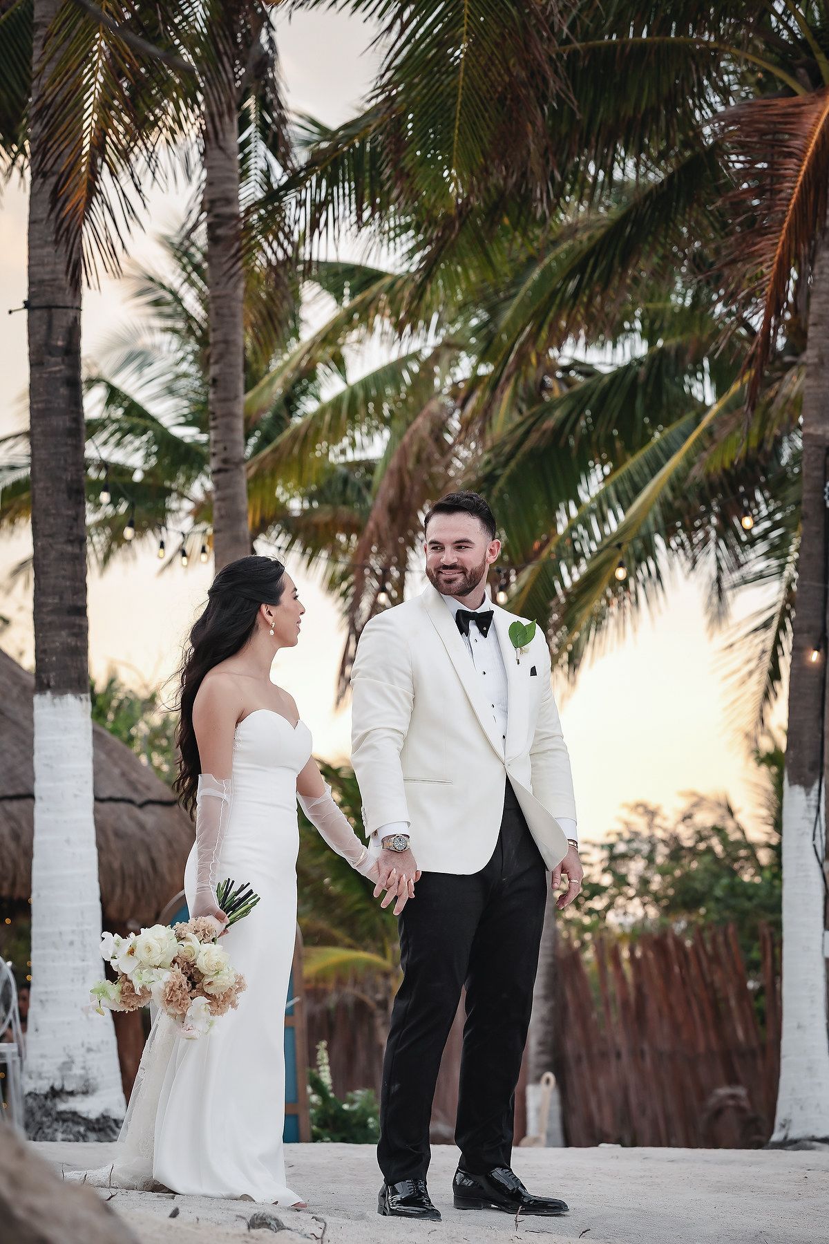 Bride and groom hold hands among tropical palm trees at sunset, she in strapless gown, he in white tuxedo jacket.