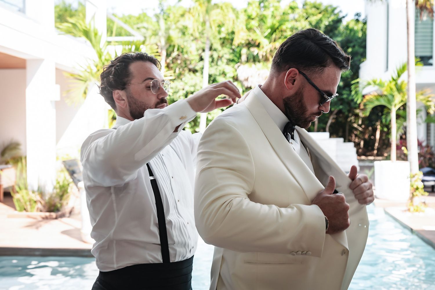 A man in a white suit being helped by another man adjusting his collar near a pool at a wedding.