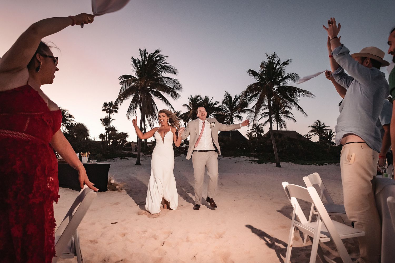 Newlywed couple walks through cheering guests on a sandy beach at sunset with palm trees in background.