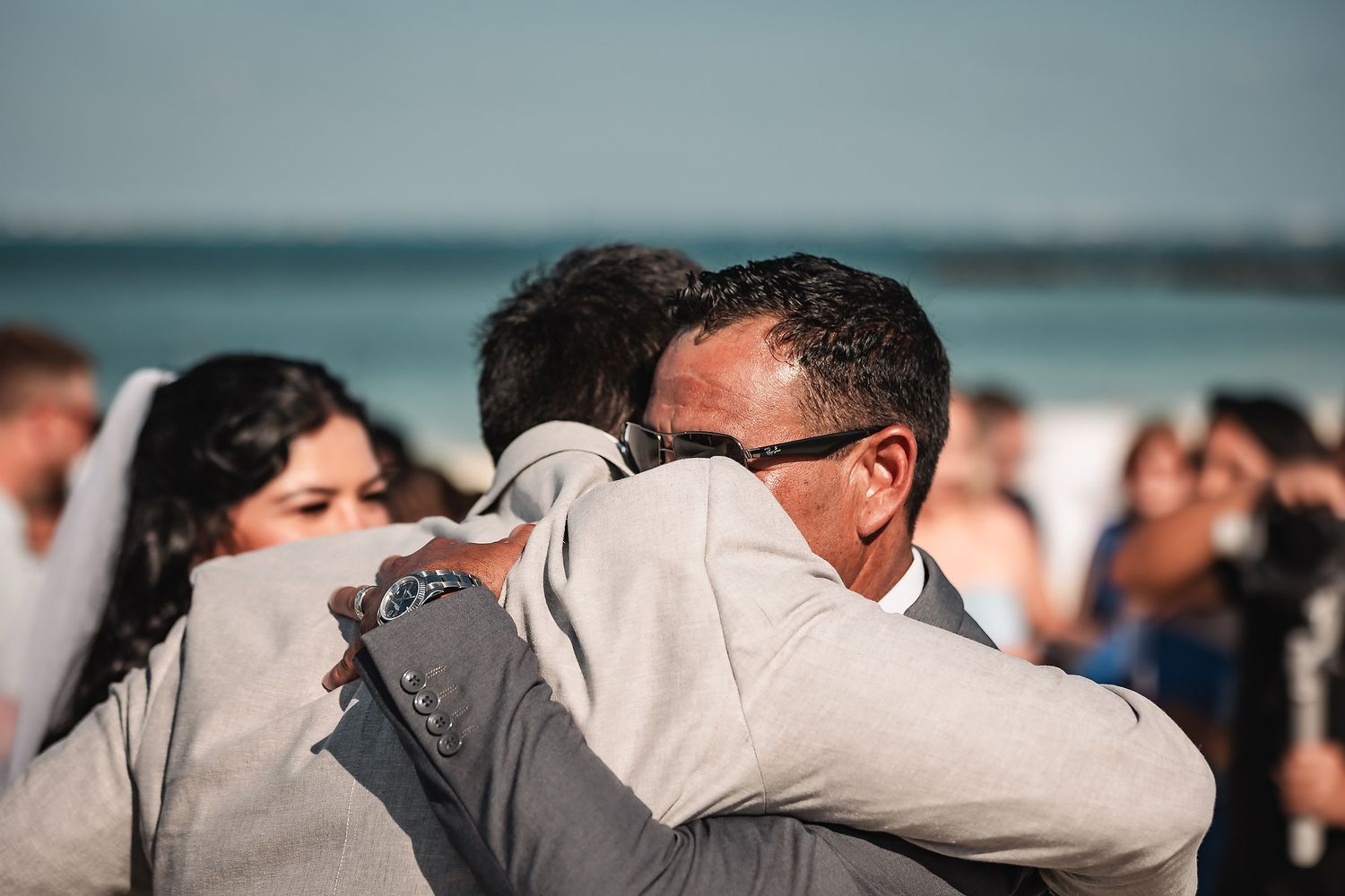 Emotional beach wedding hug between groom and guest with ocean backdrop.