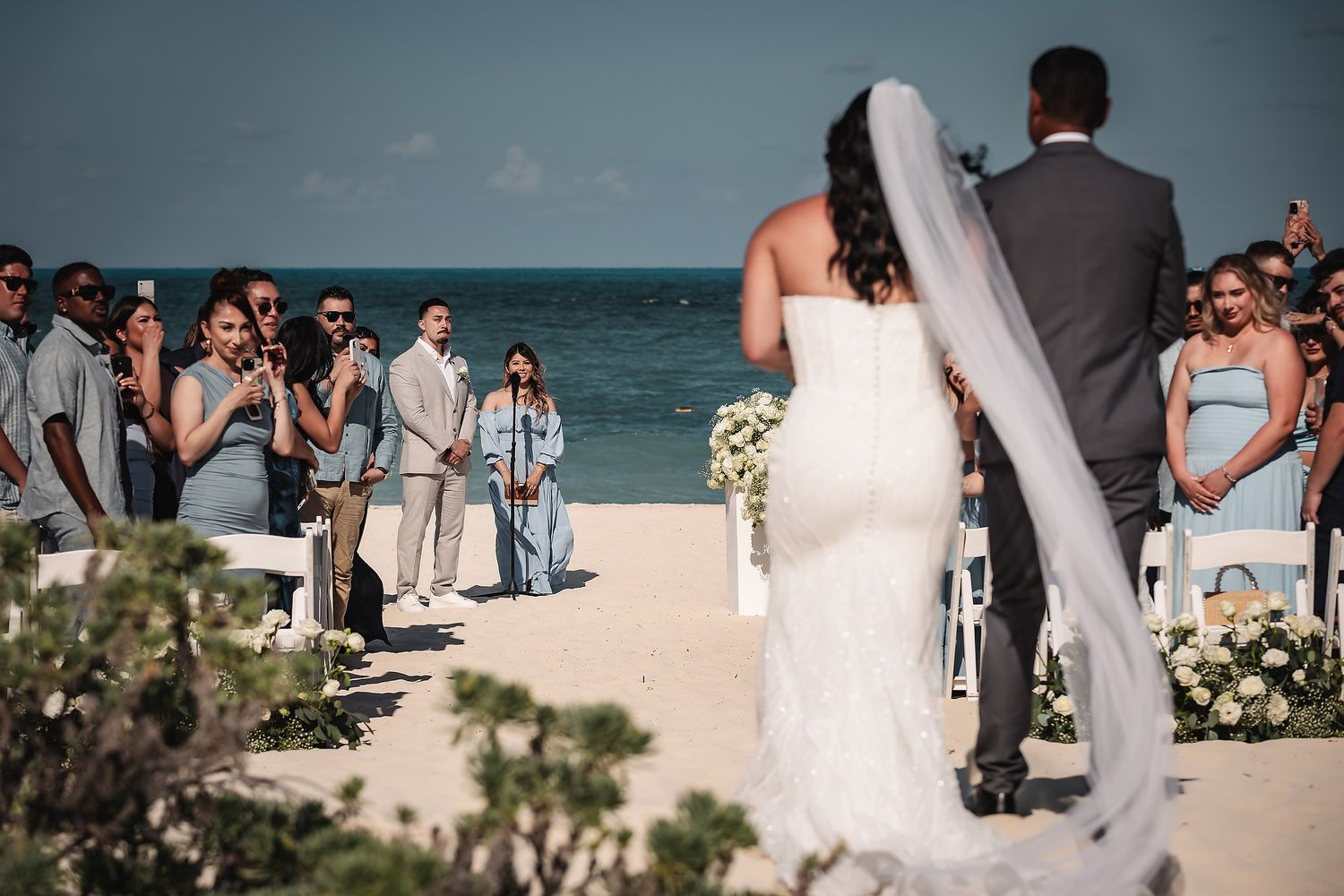 Bride and groom walk down sandy beach aisle toward officiant with ocean backdrop at outdoor destination wedding ceremony.