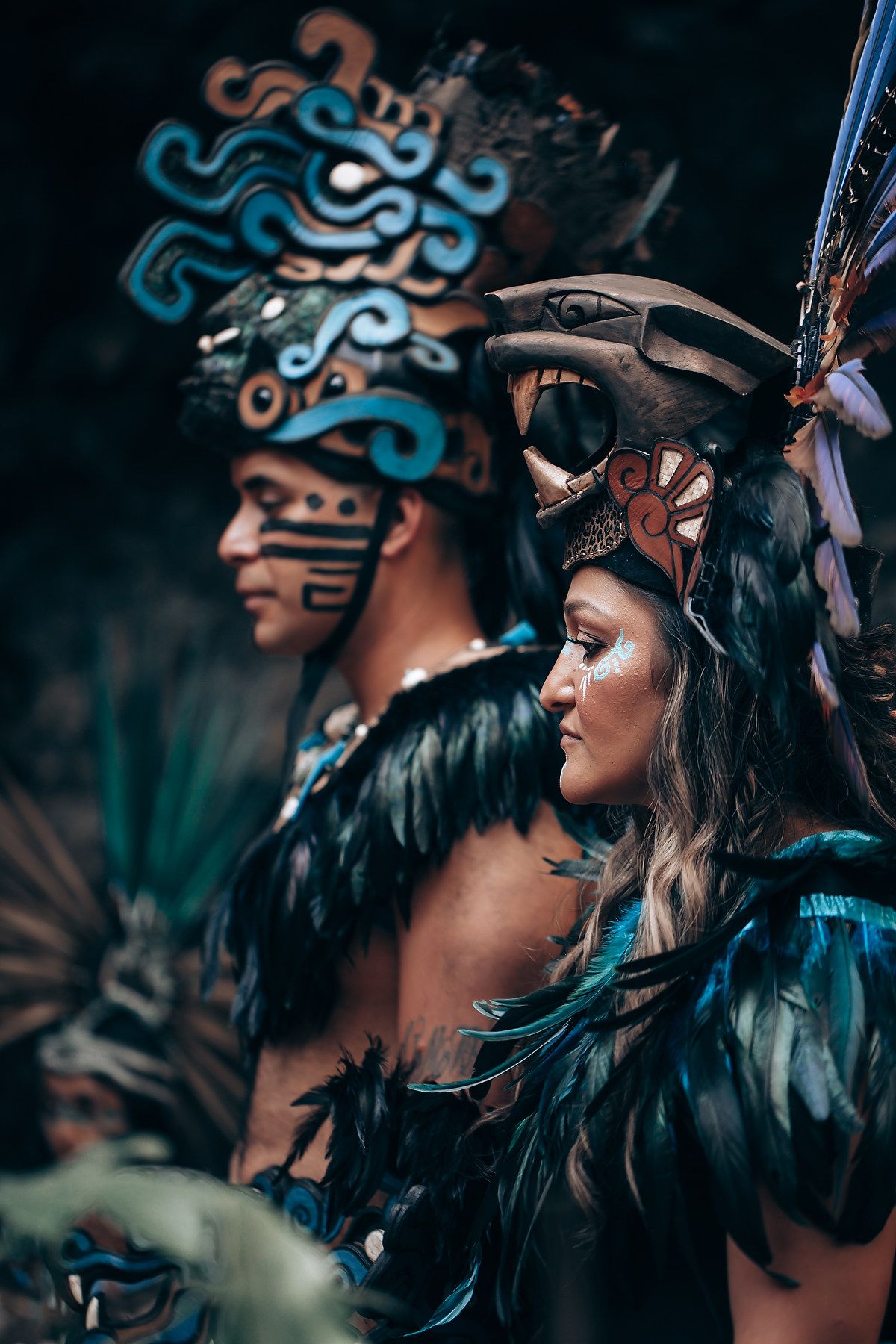 Two performers in stunning Mayan regalia with turquoise feathers and ornate headdresses pose in a dark forest backdrop.