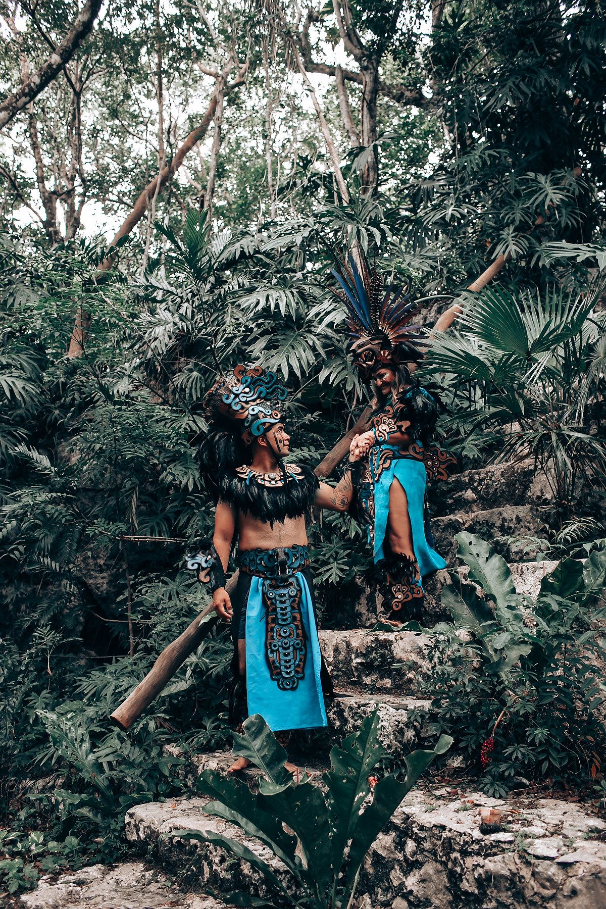 Two performers in traditional Mayan blue costumes with feathered headdresses on stone steps in a tropical jungle.